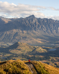 A young hiker stands at the top of a hill near Queenstown New Zealand, witnessing the beauty and awe of The Remarkables at sunset.