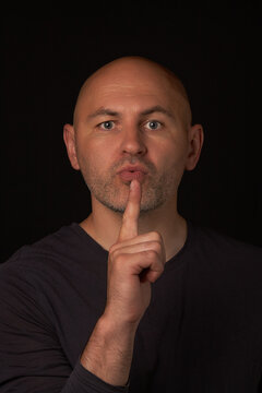 Bald unshaven man looks into the camera and blows on the index finger of his hand, on a black background. The condition of the fictional gun after the shot. Vertical picture