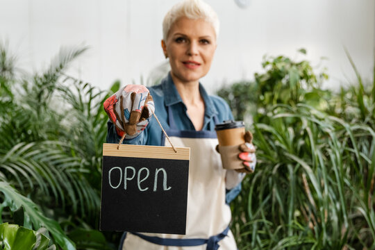 Beautiful woman just opened her flower shop. Happy woman with an open sign standind near flower shop. Happy excited woman showing OPEN sign. Attractive successful nursery owner in her greenhouse.