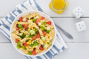 Fresh homemade colorful vegan fusilli pasta salad with beans, corn, tomato, cucumber and green bell pepper served in bowl, photographed overhead on white wood (Selective Focus, Focus on the salad)