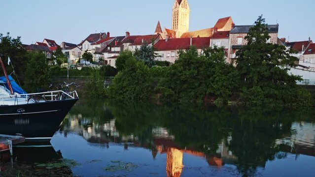 Evening view of Dormans townscape with tall lighted bell tower of Saint-Hippolyte church rising above residential areas and reflected in water of Marne river in summer, France