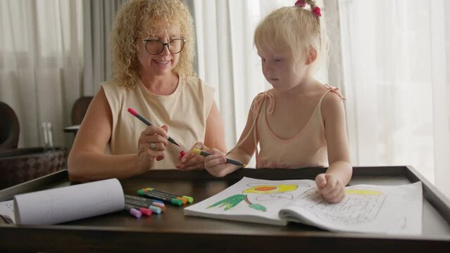 Smiling happy grandmother teaching her granddaughter how to draw with pencils in a coloring book.