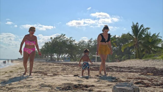 Little hispanic toddler walking along the beach with his auntie and his grandma looking for seashells on a sunny afternoon