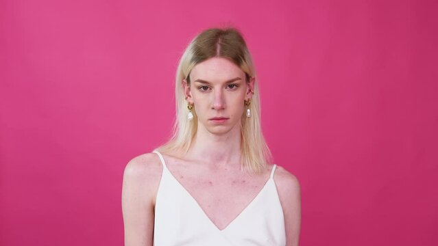 Portrait of young transgender man in a dress puts on earrings on a pink background