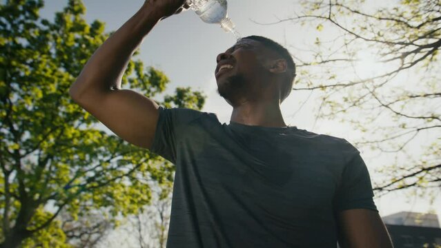 Young black man in t-shirt taking a break and drinking water by trees in park