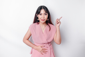 Obraz premium Starving Asian woman dressed in pink, pointing at the copy space on top of her, isolated by white background