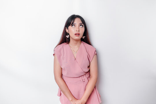 Dissatisfied young Asian woman dressed in pink blouse rolling her eyes and raising hand discontentedly, standing against white background