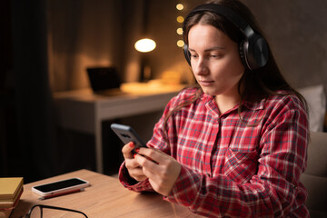 Girl student in headphones using a smartphone to listen to music or watch movies in the evening in dormitory.