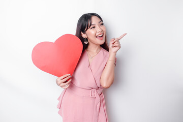 Excited Asian woman dressed in pink, pointing at the copy space beside her while holding a big red heart-shaped paper, isolated by white background