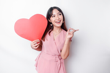 Excited Asian woman dressed in pink, pointing at the copy space beside her while holding a big red heart-shaped paper, isolated by white background