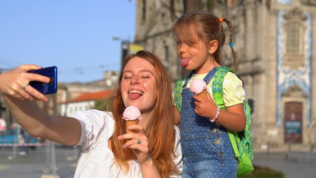 A happy young mother and little daughter with ice cream showing tongues and taking Photos On a Phone On the Street. Summertime