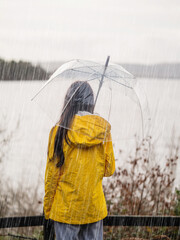 Teenager girl in yellow jacket holding translucent umbrella looking at a beautiful lake in the background in a rain. Selective focus. Outdoor activity. Going out.