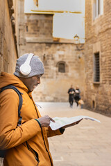 Naklejka na ściany i meble Young man on vacation with winter clothes, headphones and map visiting the gothic quarter of Barcelona (Spain).