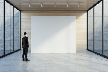 Fototapeta na wymiar Man in black suit back view looking at blank white partition with place for your logo or advertising text on light wooden background in empty hall with transparent walls and concrete floor, mock up