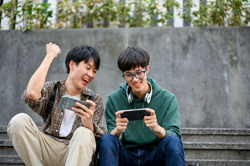 Obraz premium Cheerful Asian male college student playing mobile game with his friend, cheering his friend