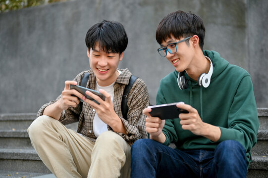 Happy young Asian man college student playing mobile game on his smartphone with his friend - Powered by Adobe