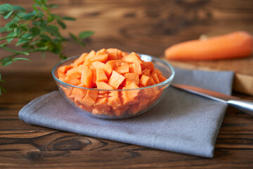 Diced carrots in a glass plate on a wooden table with a knife and whole carrots, rustic style. Diced carrots.
