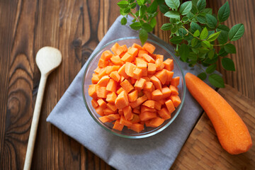 Cubes of ripe carrots in a glass bowl on a gray napkin, viewed from above on a wooden background with greenery.