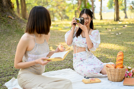 Two attractive Asian women picnic in the park together, taking a photo from retro camera