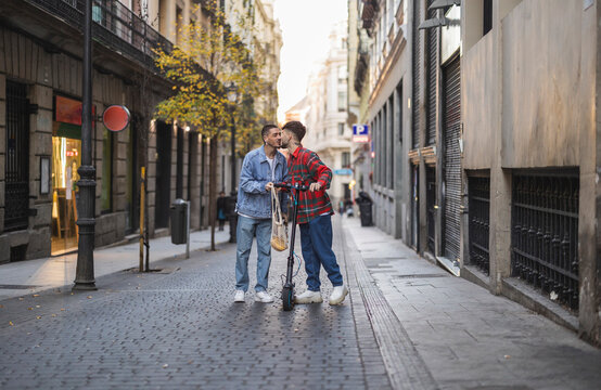 Happy gay couple kissing with electric push scooter amidst buildings