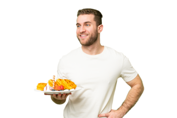 Young handsome blonde man holding waffles over isolated chroma key background posing with arms at hip and smiling