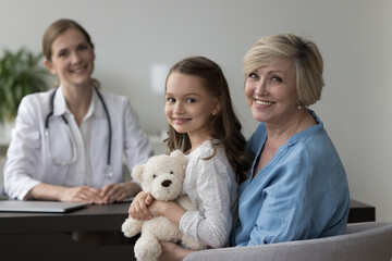 Happy older grandma holding cheerful granddaughter kid with toy in arms, looking at camera with...