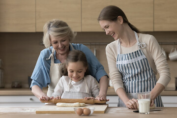 Cheerful kid girl helping granny and mom to bake in home kitchen, learning to roll, knead dough on board, smiling, laughing. Girl and women of three family generations preparing homemade bakery food