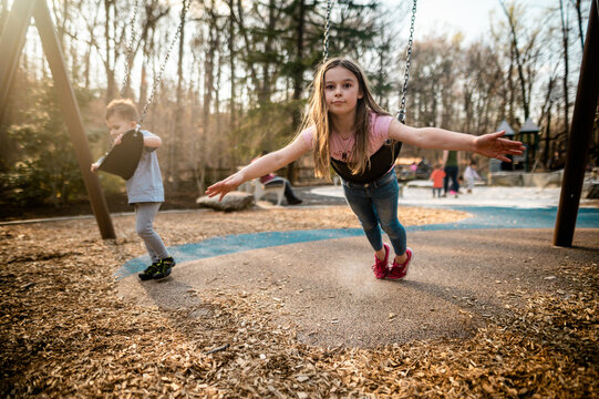 Brother and sister play on the swings at playground at sunset
