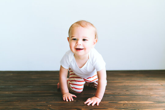 A baby boy crawls across a dark wood floor in front of a white wall