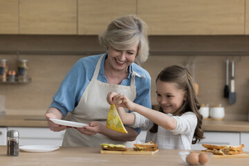 Cheerful senior granny teaching grandkid girl to prepare breakfast, snack for lunch. Happy grandma and child enjoying cooking hobby, leisure, cooking sandwiches, stacking toasts, smiling, laughing