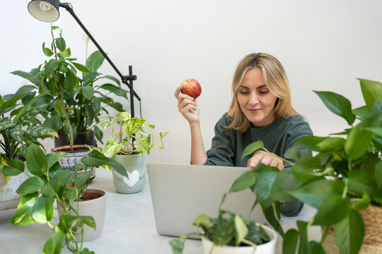 Smiling freelancer holding apple and using laptop at desk