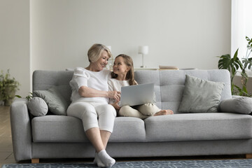 Happy granny and gen Z grandkid girl resting close on couch at home, holding laptop computer, using domestic Internet communication, online app for shopping, enjoying leisure with gadget