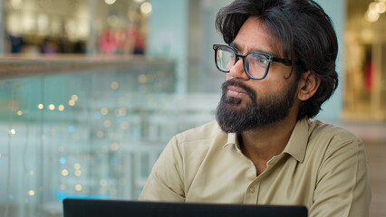 Concentrated pensive thoughtful Indian Arabian ethnic multiracial bearded businessman trader man entrepreneur male manager CEO in eyeglasses with laptop indoor looking to side thinking pondering think