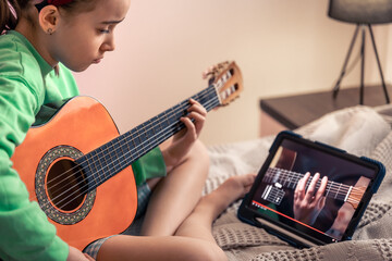 A little girl learns to play the guitar with the help of a video lesson.