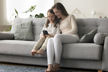 Fototapeta premium Happy little daughter kid sitting close to positive mom, holding mobile phone, using online educational app, playing video game, looking at screen, smiling, laughing, resting on home sofa