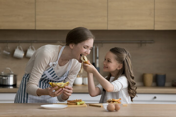 Joyful little kid girl giving mom sandwich to bite. Happy mother and daughter cooking snack for...
