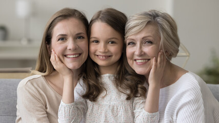 Happy sweet daughter kid touching cheeks, faces of mother and senior grandma. Little girl, mom, grandmother sitting together on home sofa, hugging, looking at camera, smiling. Family portrait