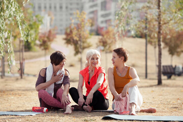 Active senior women doing yoga exercises or fitness training in park, relaxing and talking after workout. Happy Mature female friends outdoors