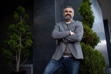 a mature satisfied male businessman is standing in a jacket at the entrance to the business center with his arms crossed on his chest