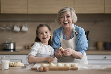 Happy excited grandma and joyful grandkid girl wearing aprons, baking at kitchen table together,...