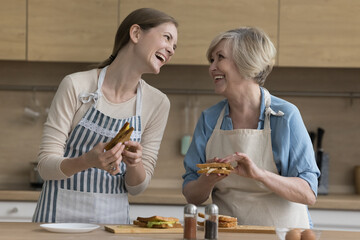 Cheerful mature mom and adult daughter woman wearing aprons, preparing sandwiches at table, cooking snacks for lunch in kitchen together, laughing, talking, chatting, enjoying culinary