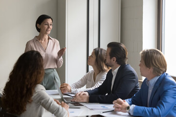 Mature female boss talking to employees on office meeting, standing at table, speaking, instructing...