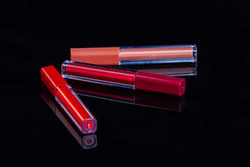 Three lip gloss sticks on reflective surface. Cosmetic makeup tools on black background.