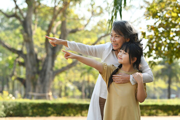 Smiling middle age grandmother and lovely granddaughter pointing to something while walking in public park at morning