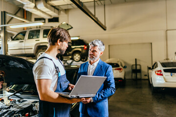 Handsome male mechanic wearing uniform, using laptop talking with grey hair business man client, finish repairing car or automobile components, working in car maintenance service center or shop.