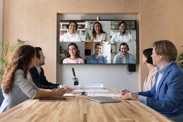 Naklejka na ściany i meble Millennial business team meeting in boardroom, talking on video conference call to happy multiethnic remote distant freelance employees, turning looks to head shots set on big screen