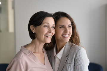 Happy beautiful senior mom and successful adult daughter standing close, hugging with face, cheek touches, looking away with dreamy toothy smiles, laughing. Family portrait