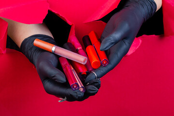 Gloved hands holding pile of various lip gloss sticks. Breaking through a red paper wall.