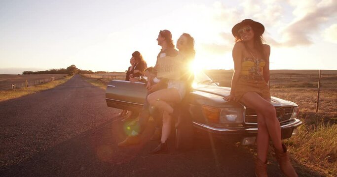 Side view of a group of teenager friends on a convertible car raising arms to celebrate a summer road trip with summer sunset sun flare