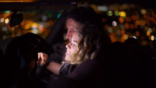 Young couple looking at pictures on a digital camera or smart phone hugging in a convertible car at night with city lights in the background